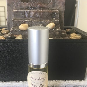 May include: A silver-capped serum bottle with a cream-colored label that reads "Theoria's Beauty Anti-Aging Serum." The bottle is filled with a yellow liquid. The background includes a small decorative fountain with water flowing.
