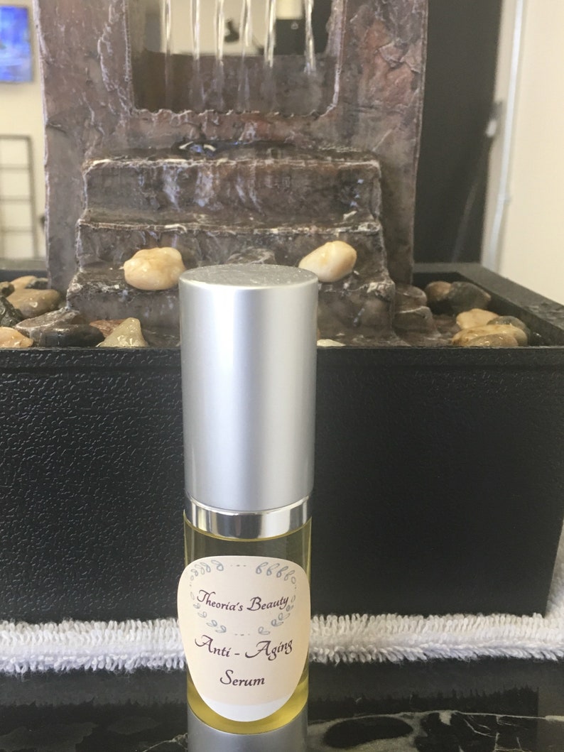 May include: A silver-capped serum bottle with a cream-colored label that reads "Theoria's Beauty Anti-Aging Serum." The bottle is filled with a yellow liquid. The background includes a small decorative fountain with water flowing.