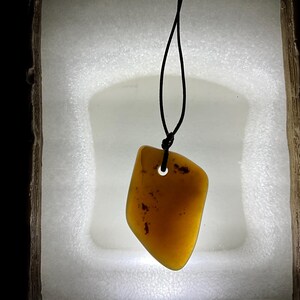May include: A yellow amber pendant with a hole in the center, hanging from a black cord. The pendant is on a white background.