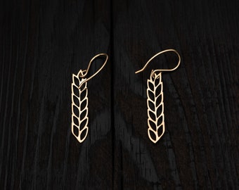 May include: A pair of gold earrings shaped like wheat stalks. The earrings are on a black wooden surface.