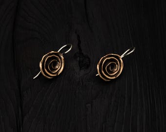 May include: A pair of bronze earrings with a rose-like design. The earrings are on a black wooden surface.