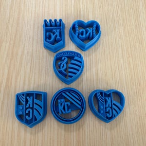 May include: Six blue plastic cookie cutters in different shapes, including a heart, a circle, and a shield. The cutters feature the letters "KC" and a baseball design.