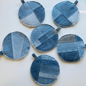 May include: Six round denim coasters with small fabric loops. Each coaster is made from patches of blue jean material in various shades of blue. The coasters are arranged on a white surface.