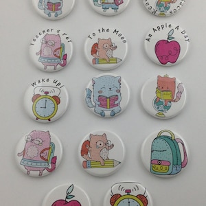 May include: A collection of white, round buttons with colorful illustrations and text. Designs include cartoon cats reading books, apples, alarm clocks, and a backpack. Text includes "#WordNerd", "Cool Cats Read", and "An Apple A Day".