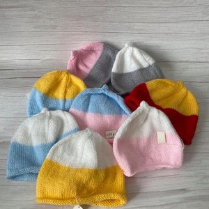 May include: A collection of eight knitted baby hats in various colour combinations, including pink, yellow, blue, red, white, and grey. The hats are arranged on a wooden surface.