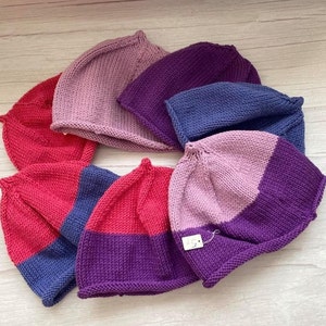 May include: Six knitted hats in various shades of pink, purple, and blue. The hats are all made with a simple stitch pattern and have a rounded crown.