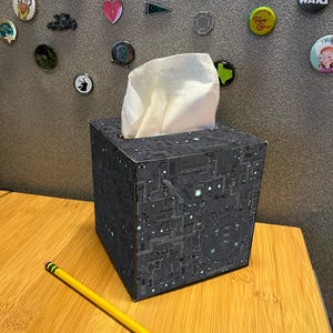 Puede incluir: Una caja de pañuelos negra con un diseño de placa de circuito, con pañuelos blancos saliendo por la parte superior. Un lápiz amarillo con una goma de borrar rosa descansa sobre una superficie de madera. Varios pines y botones están adheridos a la pared gris del fondo.