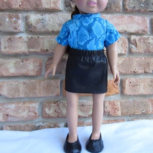 May include: A doll wearing a blue and black snake print shirt and a black skirt. The doll is standing in front of a brick wall.