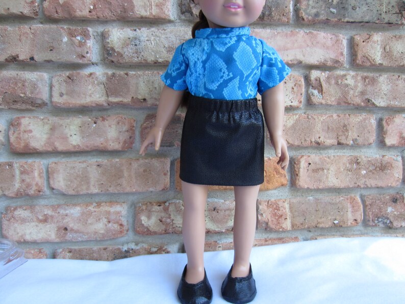 May include: A doll wearing a blue and black snake print shirt and a black skirt. The doll is standing in front of a brick wall.