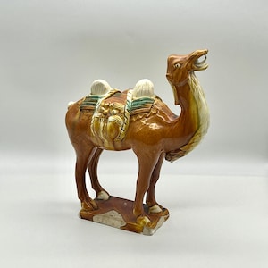May include: A ceramic camel figurine with a brown glaze and a green and white saddle. The camel is standing on a base with a brown glaze.