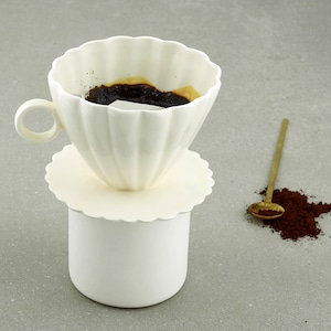 May include: White ceramic pour-over coffee maker with a white saucer and a gold spoon next to a pile of coffee grounds.