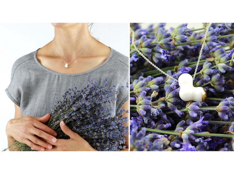 May include: A silver chain necklace with a white ceramic pendant shaped like a heart. The pendant has a gold accent on one side. The necklace is displayed on a bed of lavender flowers.