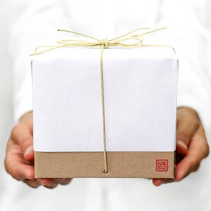 Pu&ograve; includere: Una scatola regalo bianca con fondo marrone &egrave; legata con un fiocco di spago naturale. La scatola &egrave; tenuta dalle mani di una persona.