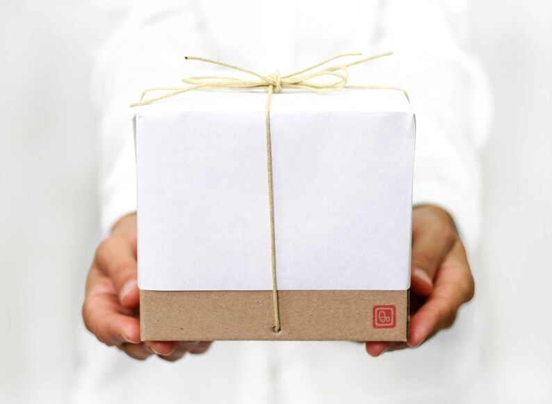 Pu&ograve; includere: Una scatola regalo bianca con fondo marrone &egrave; legata con un fiocco di spago naturale. La scatola &egrave; tenuta dalle mani di una persona.