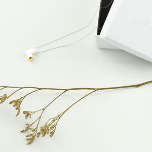 May include: A delicate silver chain necklace with a white ceramic pendant featuring a small gold accent. The necklace is displayed on a white surface with a dried flower sprig.