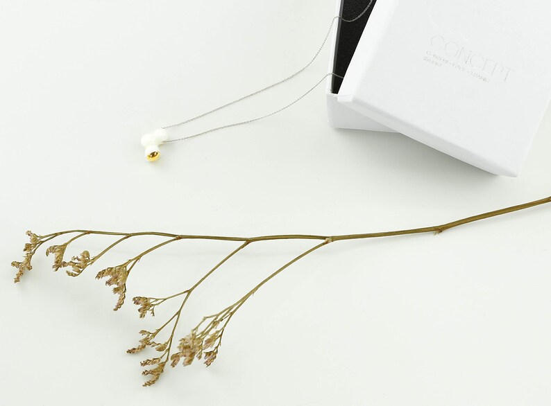 May include: A delicate silver chain necklace with a white ceramic pendant featuring a small gold accent. The necklace is displayed on a white surface with a dried flower sprig.