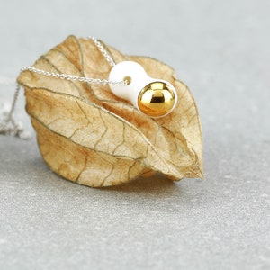 May include: A white ceramic pendant with a gold sphere on a silver chain. The pendant is shaped like a bean and has a smooth, glossy finish.