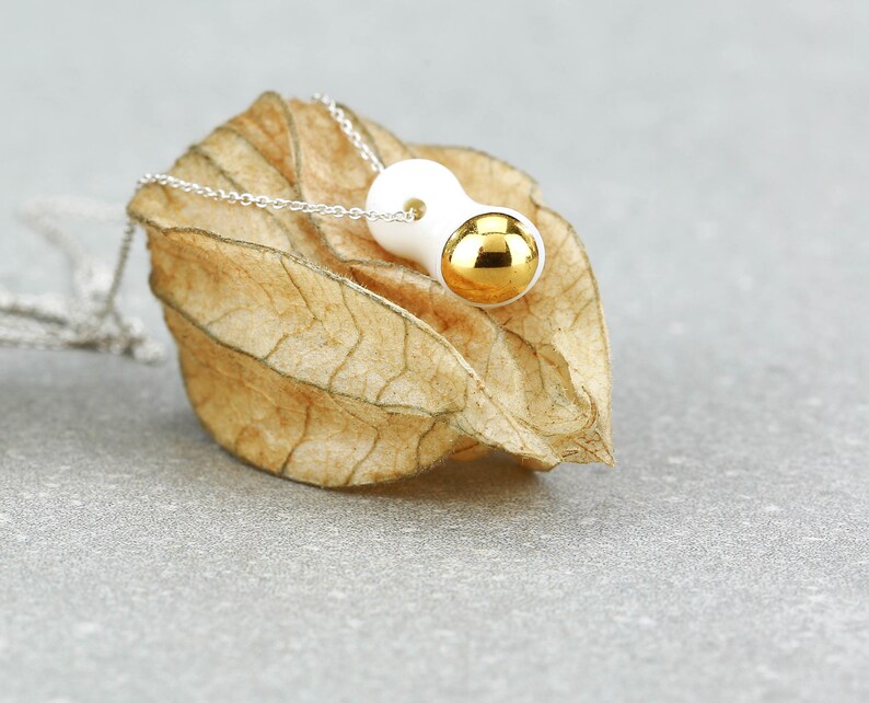 May include: A white ceramic pendant with a gold sphere on a silver chain. The pendant is shaped like a bean and has a smooth, glossy finish.