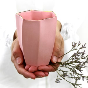May include: A pink ceramic vase with a gold rim, held in hands. The vase has a geometric shape with multiple sides.