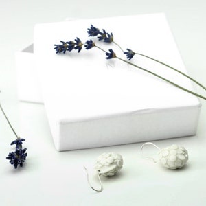 Puede incluir: Un par de pendientes de cer&aacute;mica blanca con un dise&ntilde;o texturizado y org&aacute;nico. Los pendientes cuelgan de ganchos plateados y se muestran sobre una superficie blanca con flores de lavanda secas.