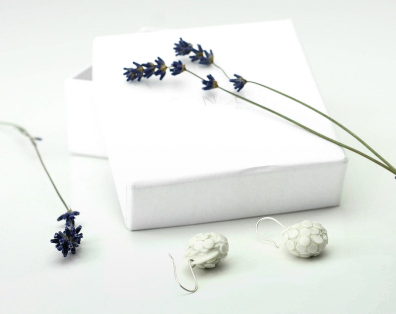 Puede incluir: Un par de pendientes de cer&aacute;mica blanca con un dise&ntilde;o texturizado y org&aacute;nico. Los pendientes cuelgan de ganchos plateados y se muestran sobre una superficie blanca con flores de lavanda secas.