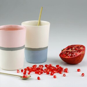 Pu&ograve; includere: Due tazze in ceramica con una striscia grigia e una striscia colorata. La tazza a sinistra &egrave; rosa e la tazza a destra &egrave; blu. Una met&agrave; di melograno e semi di melograno sono sparsi su una superficie bianca.