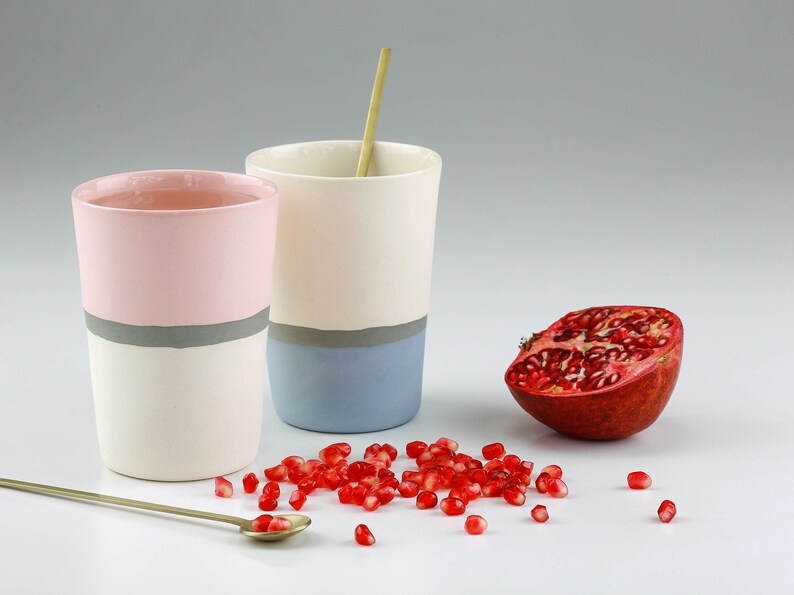 Pu&ograve; includere: Due tazze in ceramica con una striscia grigia e una striscia colorata. La tazza a sinistra &egrave; rosa e la tazza a destra &egrave; blu. Una met&agrave; di melograno e semi di melograno sono sparsi su una superficie bianca.