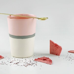 Pu&ograve; includere: Una tazza in ceramica rosa e bianca con una striscia grigia, un cucchiaio d'oro e pezzi di cioccolato rosa su una superficie bianca.