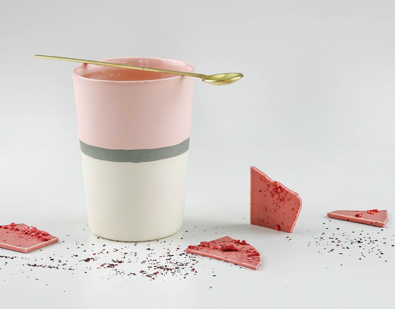 Pu&ograve; includere: Una tazza in ceramica rosa e bianca con una striscia grigia, un cucchiaio d'oro e pezzi di cioccolato rosa su una superficie bianca.