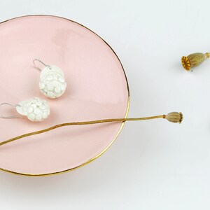 Puede incluir: Un par de pendientes de cer&aacute;mica blanca con un dise&ntilde;o texturizado con forma de flor. Los pendientes se muestran en un plato rosa con un borde dorado.