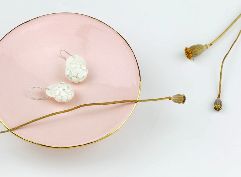 Puede incluir: Un par de pendientes de cer&aacute;mica blanca con un dise&ntilde;o texturizado con forma de flor. Los pendientes se muestran en un plato rosa con un borde dorado.