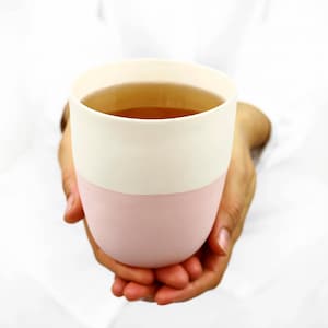May include: A pink and white ceramic mug with a handle, filled with a light brown liquid. The mug is being held by two hands.