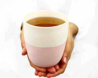 May include: A pink and white ceramic mug with a handle, filled with a light brown liquid. The mug is being held by two hands.