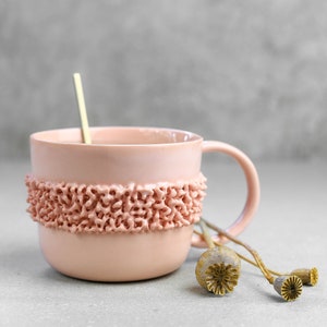 May include: A pink ceramic mug with a textured band around the middle. The mug has a handle and is sitting on a grey surface with dried poppy seed pods.