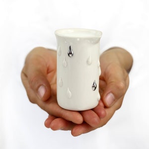 May include: A white ceramic creamer with silver raindrop accents. The creamer is being held in a person's hands.