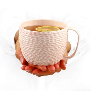 May include: A pink ceramic mug with a textured surface, filled with tea and a lemon slice. The mug is being held by a person's hands.