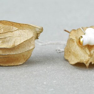 May include: A white ceramic necklace with gold accents hangs on a dried, brown, papery husk. The necklace features three rounded, organic shapes.