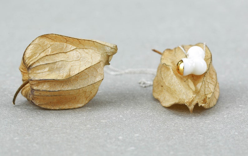May include: A white ceramic necklace with gold accents hangs on a dried, brown, papery husk. The necklace features three rounded, organic shapes.