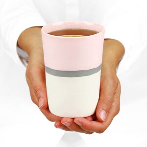 Pu&ograve; includere: Una tazza in ceramica rosa e bianca con una striscia grigia intorno al centro. La tazza &egrave; piena di un liquido marrone chiaro.