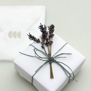 Puede incluir: Una caja de regalo blanca atada con una cuerda gris y decorada con una rama de lavanda seca.
