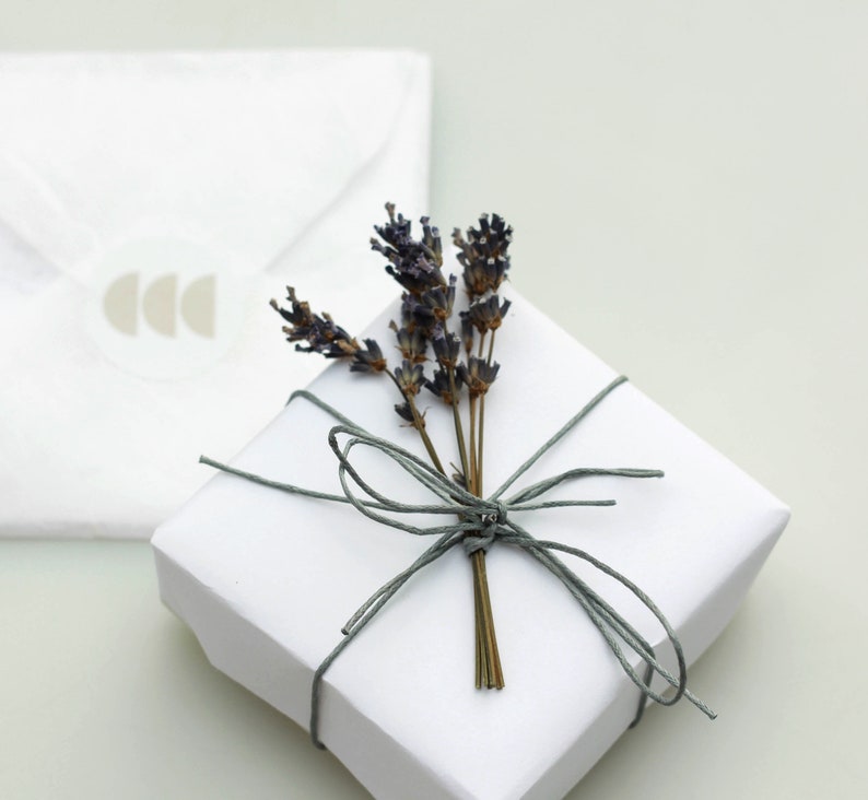 Puede incluir: Una caja de regalo blanca atada con una cuerda gris y decorada con una rama de lavanda seca.