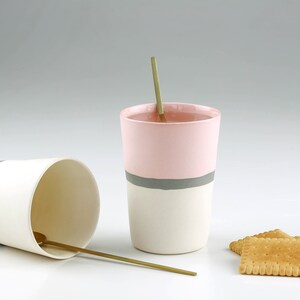 Pu&ograve; includere: Due tazze in ceramica con base bianca e una fascia colorata intorno al centro. La tazza a sinistra ha una fascia blu chiaro e la tazza a destra ha una fascia rosa con una striscia grigia. Entrambe le tazze hanno un cucchiaio dorato all'interno. Ci sono tre cracker marroni su una superficie bianca.