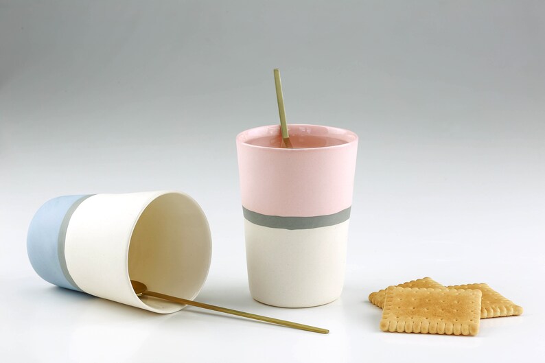 Pu&ograve; includere: Due tazze in ceramica con base bianca e una fascia colorata intorno al centro. La tazza a sinistra ha una fascia blu chiaro e la tazza a destra ha una fascia rosa con una striscia grigia. Entrambe le tazze hanno un cucchiaio dorato all'interno. Ci sono tre cracker marroni su una superficie bianca.
