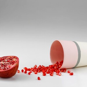 Pu&ograve; includere: Una met&agrave; di melograno con semi rossi che si riversano da una tazza di ceramica a strisce rosa e bianche.