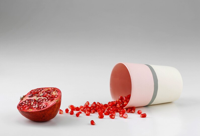 Pu&ograve; includere: Una met&agrave; di melograno con semi rossi che si riversano da una tazza di ceramica a strisce rosa e bianche.