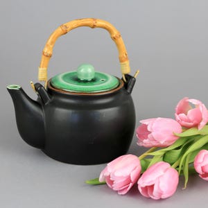 Op de afbeelding: Een zwarte keramische theepot met een bamboe handvat en een groen deksel. De theepot wordt vergezeld door een boeket roze tulpen. De handgreep van de theepot is van bamboe.