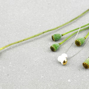 May include: A white ceramic pendant with a gold accent hangs on a silver chain. The chain is draped over dried poppy seed pods.