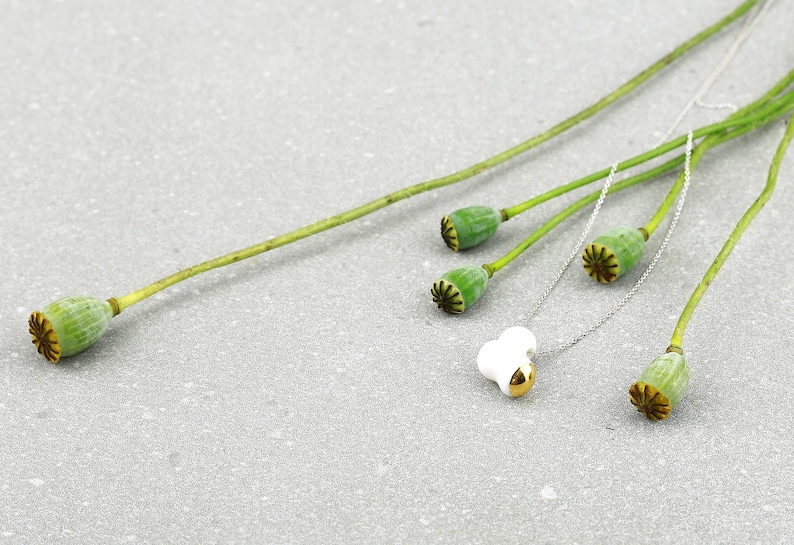 May include: A white ceramic pendant with a gold accent hangs on a silver chain. The chain is draped over dried poppy seed pods.