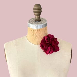 May include: A deep burgundy fabric scrunchie with a gathered, ruffled texture. The hair accessory is displayed on a beige, vintage-style dress form mannequin that features a metallic neck piece and visible vertical seam lines on the torso.