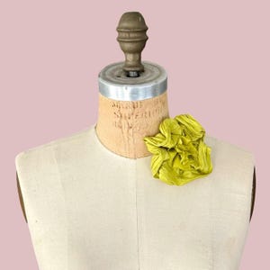 May include: A chartreuse fabric flower brooch with layered, ruffled petals attached to a beige canvas dress form. The dress form features a metallic neck piece and a visible printed label on the upper chest area, displaying a textured surface.